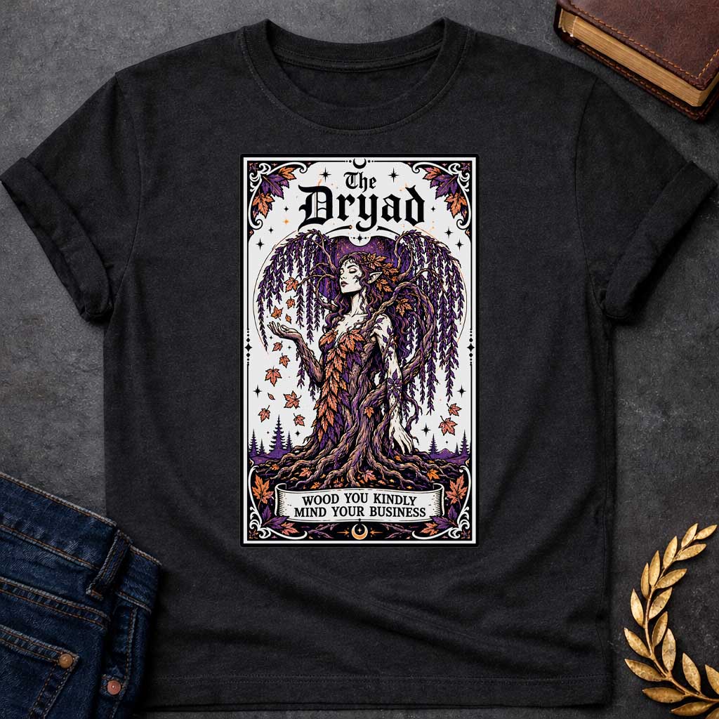 Black T-shirt with a mystical, tarot-card style design framed by an ornate border. It features a majestic weeping willow dryad whose gown is woven from purple and orange autumn leaves. At the top, bold gothic text reads "The Dryad." A weathered banner at the base of her rooted trunk features a clever, pun-based tagline that reads: "WOOD YOU KINDLY MIND YOUR BUSINESS" in a clean, black serif font