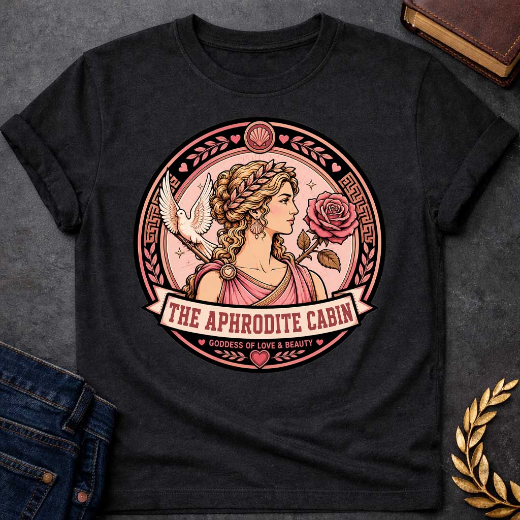 Black t-shirt with a Aphrodite Greek mythology cabin design featuring the goddess of love and beauty in a pink robe and laurel crown, surrounded by a rose, white dove, hearts, seashells, and Greek key details. Circular pink and black emblem includes the text “The Aphrodite Cabin” and “Goddess of Love & Beauty,”