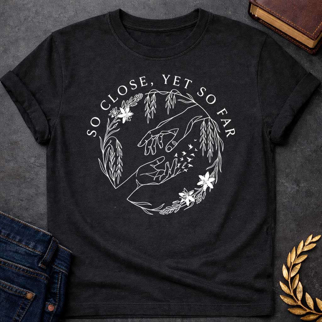 Black t-shirt in delicate fine-line style: two reaching hands almost touch, with one hand dissolving into a flock of small birds mid-fingertip, evoking Orpheus and Eurydice. Willow branches and small flowers form a circular wreath around the scene, with elegant serif text arcing above