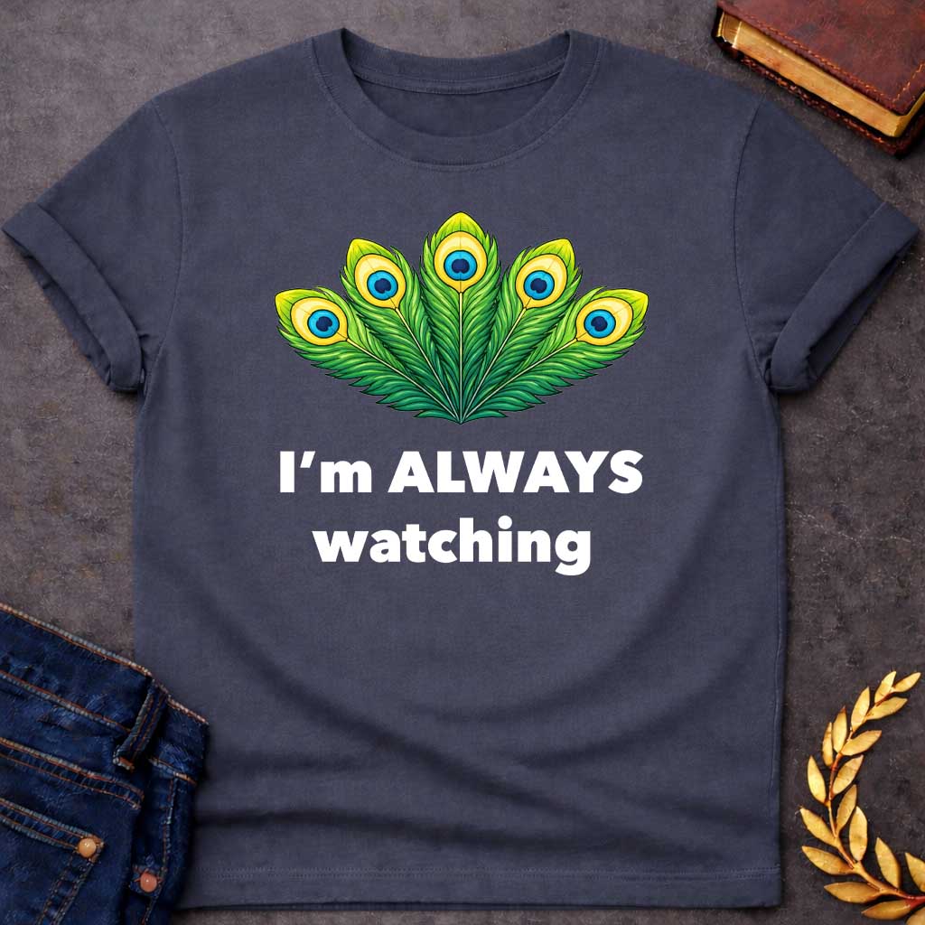 Dark Heather T-shirt graphic featuring a vibrant illustrated fan of five peacock feathers in rich green, yellow, and blue tones, each with a distinctive eye-spot, above text reading "I'm Always Watching" — a humorous reference to Hera, the Greek Queen of the Gods, whose sacred symbol is the peacock