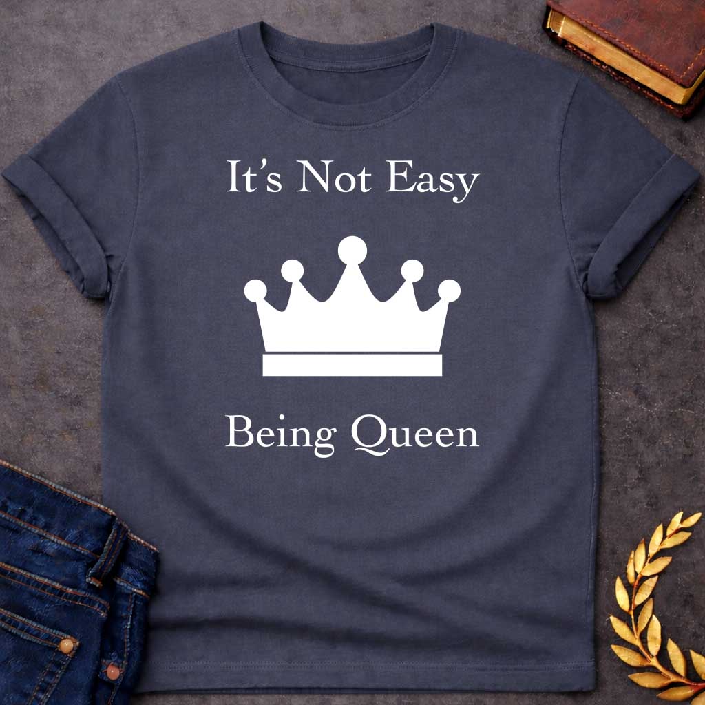 Dark Heather T-shirt graphic in white on black featuring elegant serif text reading "It's Not Easy Being Queen" split above and below a simple white crown silhouette icon, in a humorous nod to Hera, the Greek Queen of the Gods