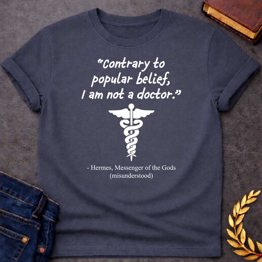 Dark Heather T-shirt graphic in white text featuring a handwritten-style quote reading "Contrary to popular belief, I am not a doctor." above a white caduceus symbol, with the humorous attribution below in serif text reading "- Hermes, Messenger of the Gods (misunderstood)," playing on the common confusion between Hermes' caduceus and the medical symbol
