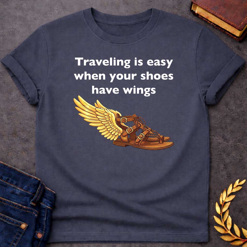 Dark Heather T-shirt graphic featuring bold white text reading "Traveling is easy when your shoes have wings" above a detailed illustration of Hermes' winged sandal, depicted as a brown leather gladiator sandal with gold buckles and braided lacing, with a large golden wing extending from the ankle — a humorous reference to the talaria, the iconic flying sandals of Hermes, the Greek Messenger God