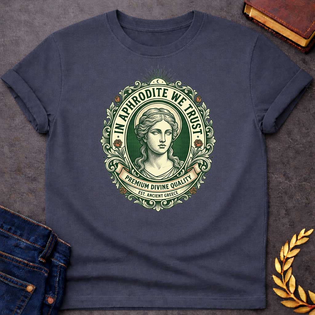Dark Heather T-shirt graphic featuring a vintage apothecary label-style oval badge design in forest green, cream, and terracotta. A detailed engraving-style portrait of Aphrodite is centered within the badge, surrounded by ornate scrollwork and flanked by red roses, with a crescent moon and sunburst radiating from the top. Text arched around the inner circle reads "In Aphrodite We Trust," with a scroll banner below reading "Premium Divine Quality" and smaller text underneath reading "Est. Ancient Greece,"