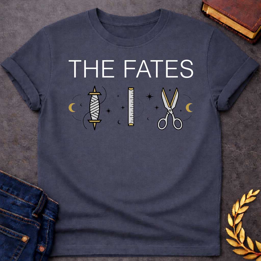 Dark Heather T-shirt graphic featuring large bold white text reading "The Fates" above three minimalist icons representing the tools of the Moirai, the Greek Fates — a spindle with thread, a measuring ruler, and a pair of scissors — each surrounded by subtle crescent moons and celestial star details, rendered in white and gold tones