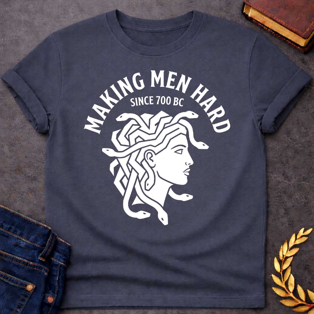 Dark Heather T-shirt graphic white featuring a bold flat graphic side-profile illustration of Medusa with snake hair, in a minimalist ancient Greek coin or seal style. Bold arched text above reads "Making Men Hard" with smaller text below it reading "Since 700 BC" — a witty double entendre referencing Medusa's mythological power to turn men to stone