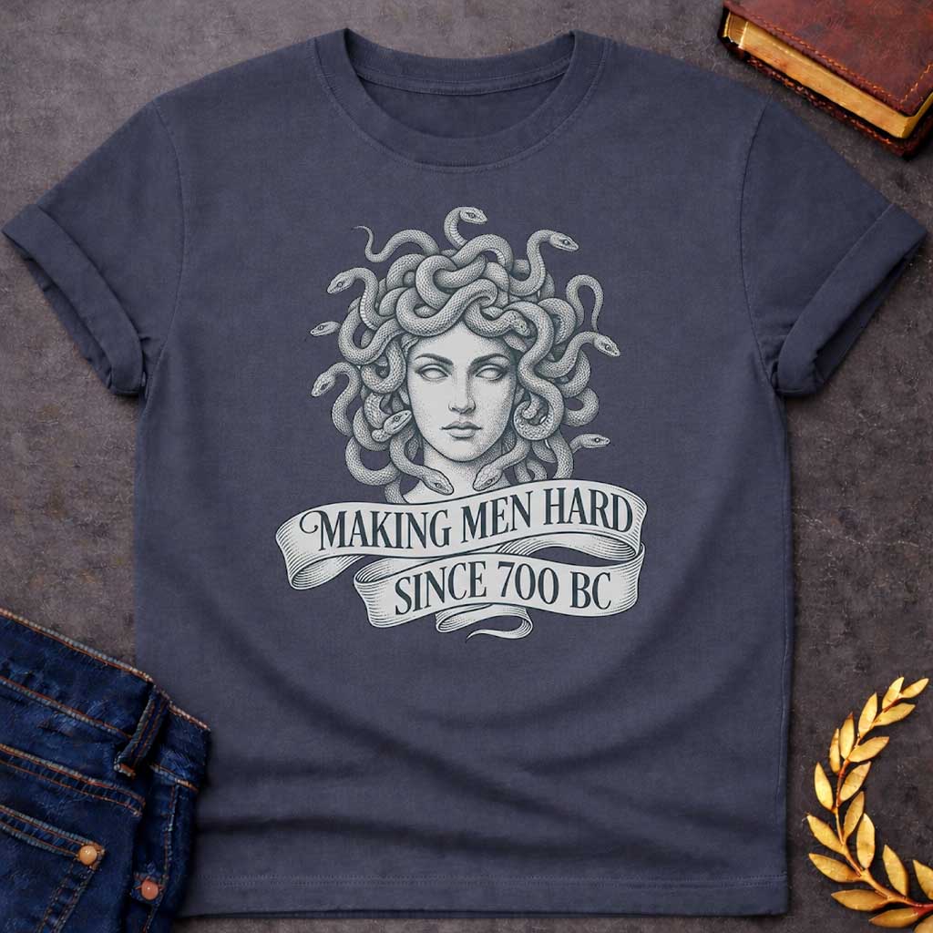 Dark Heather T-shirt graphic in grey and white featuring a highly detailed fine-line tattoo-style front-facing portrait of Medusa with numerous coiling snakes for hair, rendered in intricate stippling and engraving technique, with a flowing ribbon banner below reading "Making Men Hard Since 700 BC" — a witty double entendre referencing Medusa's mythological power to turn men to stone