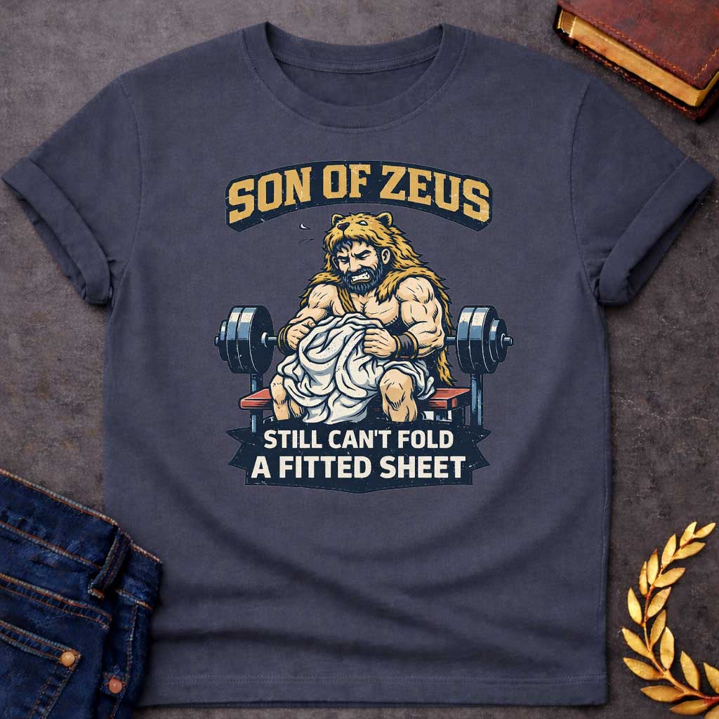 Dark Heather T-shirt featuring a humorous illustration of a muscular, bearded man wearing a lion pelt—resembling Hercules—sitting on a weight bench. He is struggling, with a frustrated expression, to fold a crumpled white fitted sheet. Behind him is a barbell. The text at the top reads "SON OF ZEUS" in a distressed, arched font, and the text at the bottom reads "STILL CAN'T FOLD A FITTED SHEET" in a banner