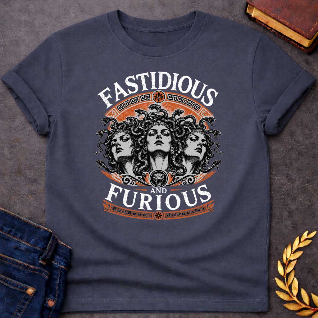 Dark Heather T-shirt with a Humorous design combining 'Fast and Furious' with Greek mythology. Text reads 'FASTIDIOUS AND FURIOUS' in distressed lettering surrounding three faces of Medusa in black and white engraving style. Detailed serpents with individual scales form her hair. Greek key pattern borders in orange frame the composition with decorative sun motifs and a small snake head icon at center. Blends classical mythology with contemporary pop culture humor and clever wordplay.