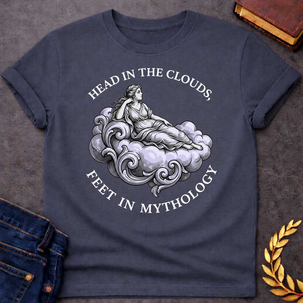 Dark Heather T-shirt with a classical Greek mythology t-shirt design featuring a detailed engraving-style illustration. A woman in a traditional draped chiton relaxes gracefully, reclining on a large, swirling baroque-style cloud shaded in a pale purple hue. Arched above her is the text "HEAD IN THE CLOUDS," and curved inversely below the cloud is "FEET IN MYTHOLOGY," all written in a clean white serif font.