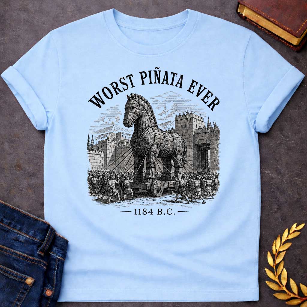 Light Blue T-Shirt with a detailed, woodcut-style graphic design. The central illustration depicts the wooden Trojan Horse being pulled on a wheeled platform by many figures using ropes toward the stone gates of a walled city. Arched over the top of the scene in a distressed, black serif font is the text "WORST PIÑATA EVER." At the bottom of the design, the year "1184 B.C." is printed between two horizontal decorative dashes