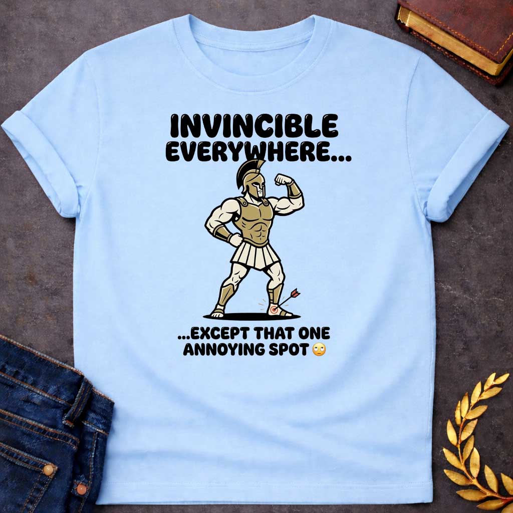 Light Blue T-Shirt with a humorous cartoon of a muscular Greek warrior in a bodybuilder pose, flexing his bicep. Wears gold Corinthian helmet, chest armor, white skirt, and greaves with exaggerated muscles. An arrow with target symbol has struck his heel. Text reads 'INVINCIBLE EVERYWHERE...' above and '...EXCEPT THAT ONE ANNOYING SPOT' with eye-roll emoji below. Comedic reference to Achilles' famous vulnerability—his heel was his only weak point. Cartoon style and modern slang create a lighthearted Greek m