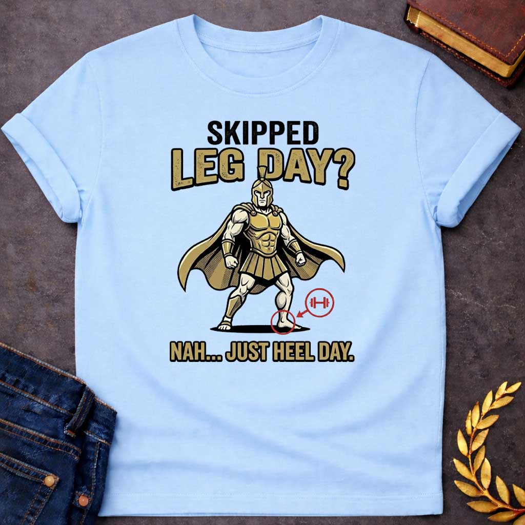 Light Blue T-Shirt with a humorous cartoon combining Greek mythology with gym culture. A muscular Greek warrior in heroic stance wears gold Corinthian helmet, armor with defined abs, pleated skirt, and cape. The warrior has an exaggerated upper body but the left heel is circled in red with an arrow pointing to a barbell icon. Text reads 'SKIPPED LEG DAY?' above and 'NAH... JUST HEEL DAY' below. Comedic mashup referencing Achilles' vulnerable heel and the modern fitness joke about skipping leg day.