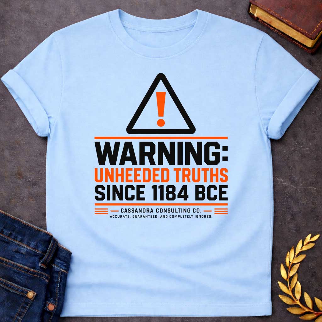 Light Blue T-shirt with a corporate-style satirical design mimicking an industrial hazard sign. At the top is a triangular warning icon with an orange exclamation point. Bold block text reads "WARNING: UNHEEDED TRUTHS SINCE 1184 BCE" in alternating black and orange. Below, it features a faux company name: "CASSANDRA CONSULTING CO." followed by the tagline, "ACCURATE, GUARANTEED, AND COMPLETELY IGNORED."