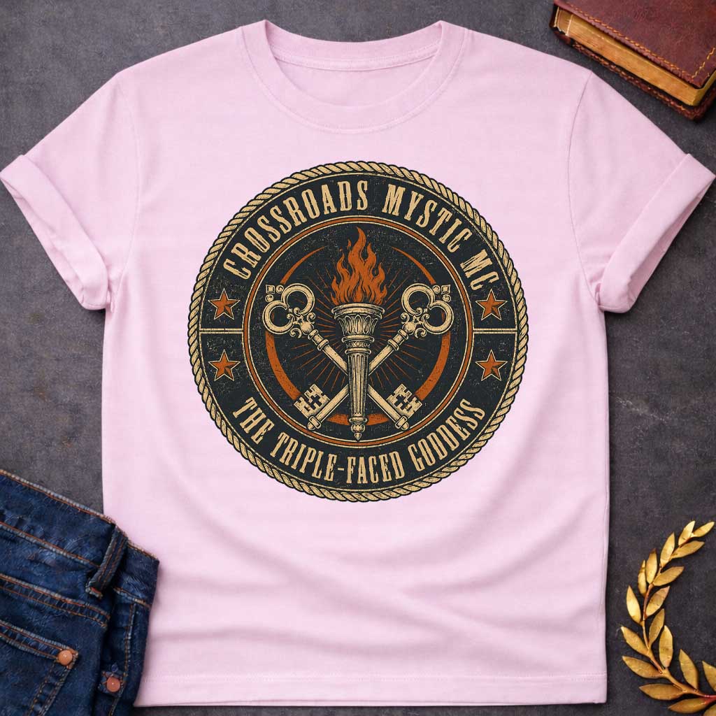 Light Pink T-shirt with a Motorcycle club patch-style emblem in navy, gold, and orange inspired by Hecate and crossroads mythology. A central ornate torch with orange flames is flanked by two crossed serpent handles with decorative gold scrollwork. Radiating lines suggest divine power. Text reads 'Crossroads Mystic MC' and 'The Triple-Faced Goddess' around the border. Orange stars and rope border complete the official club patch look. Distressed texture adds authentic biker aesthetic.