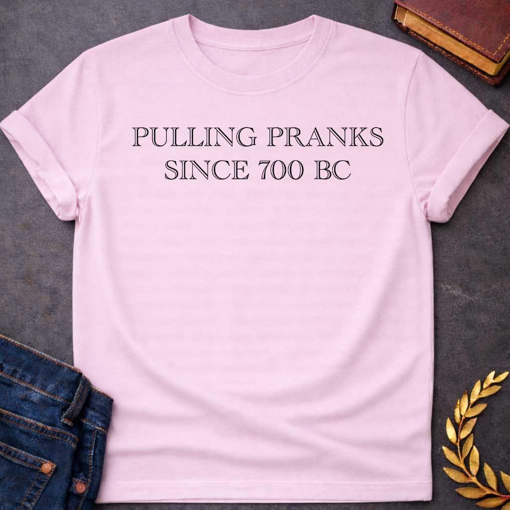 Light Pink T-shirt graphic in black text featuring large outlined engraved-style serif text reading "Pulling Pranks Since 700 BC" — a humorous nod to Hermes, the Greek Messenger God and notorious trickster