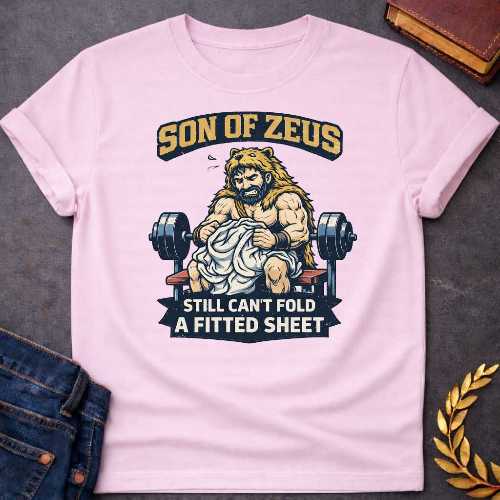 Light Pink T-shirt featuring a humorous illustration of a muscular, bearded man wearing a lion pelt—resembling Hercules—sitting on a weight bench. He is struggling, with a frustrated expression, to fold a crumpled white fitted sheet. Behind him is a barbell. The text at the top reads "SON OF ZEUS" in a distressed, arched font, and the text at the bottom reads "STILL CAN'T FOLD A FITTED SHEET" in a banner