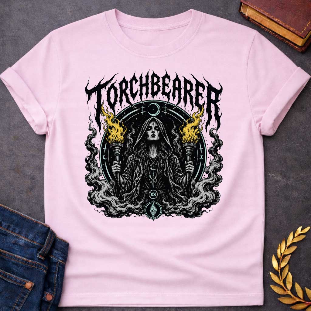 Light Pink T-shirt with a Dark metal band aesthetic illustration titled 'Torchbearer.' Hecate holds two ornate torches with gold and black flames. She wears mystical symbols including a crescent moon necklace and a circular mandala halo with occult iconography. Swirling smoke and flame designs surround the figure in black, white, and gold. Blends heavy metal imagery with Greek mythology and mystical symbolism celebrating enlightenment, divine knowledge, and dark power.