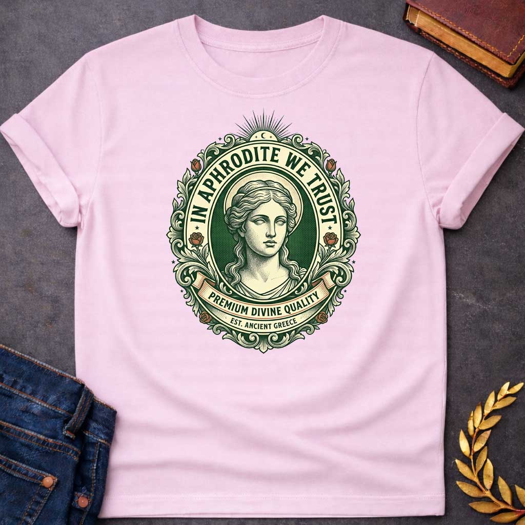 Light Pink T-shirt graphic featuring a vintage apothecary label-style oval badge design in forest green, cream, and terracotta. A detailed engraving-style portrait of Aphrodite is centered within the badge, surrounded by ornate scrollwork and flanked by red roses, with a crescent moon and sunburst radiating from the top. Text arched around the inner circle reads "In Aphrodite We Trust," with a scroll banner below reading "Premium Divine Quality" and smaller text underneath reading "Est. Ancient Greece,"