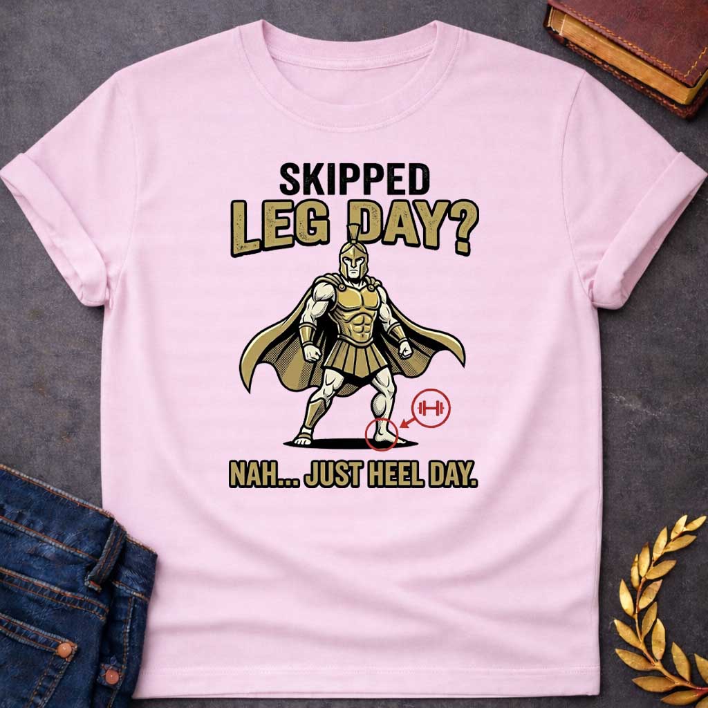 Light Pink T-Shirt with a humorous cartoon combining Greek mythology with gym culture. A muscular Greek warrior in heroic stance wears gold Corinthian helmet, armor with defined abs, pleated skirt, and cape. The warrior has an exaggerated upper body but the left heel is circled in red with an arrow pointing to a barbell icon. Text reads 'SKIPPED LEG DAY?' above and 'NAH... JUST HEEL DAY' below. Comedic mashup referencing Achilles' vulnerable heel and the modern fitness joke about skipping leg day.