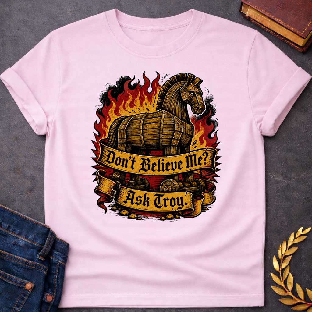 Light Pink T-shirt with a bold illustration of the Trojan Horse engulfed in flames with red, orange, and yellow fire and black smoke. The wooden horse emerges from flames with detailed wood-grain texture. Two ornate gold banners with blackletter text read 'Don't Believe Me?' and 'Ask Troy.' Dark red background with rubble scattered below. References Cassandra's ignored warning about the Trojan Horse that led to Troy's destruction. Dramatic tattoo-inspired style with vintage engraving detail.