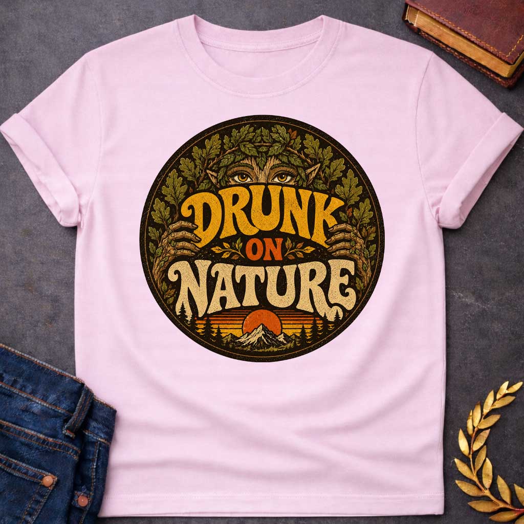 Light Pink T-shirt with a vintage, retro-style t-shirt graphic enclosed in a circular badge. At the top, the intense gold eyes of a forest nymph or dryad peek out from behind a thick crown of oak leaves. Earthy, stylized 1970s block text proudly reads "DRUNK ON NATURE" in gold and cream. The bottom of the design showcases a serene mountain landscape and a setting sun, framed by rustic pine trees and a distressed texture
