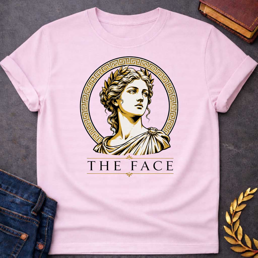 Light Pink T-shirt with a classical Greek graphic featuring a gold and cream vector portrait of Helen of Troy in profile. She wears a golden laurel wreath and a traditional draped chiton, looking upward with a serene expression. The portrait is framed by a circular border with a gold Greek key (meander) pattern. Below the emblem, the text "THE FACE" is written in a clean, elegant black serif font between horizontal gold accent lines.