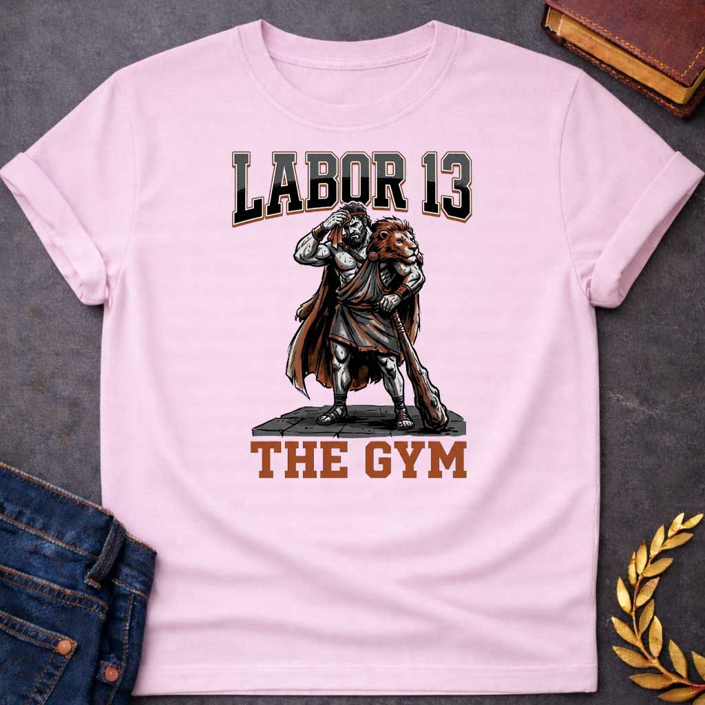 Light Pink T-shirt featuring a sweat-drenched, muscular figure resembling Heracles, wearing a lion-skin cloak and holding a large wooden club. He stands on a stone platform while wiping his brow with a towel. The text "LABOR 13" is printed in large, bold, block letters across the top, and "THE GYM" is in a matching style at the bottom