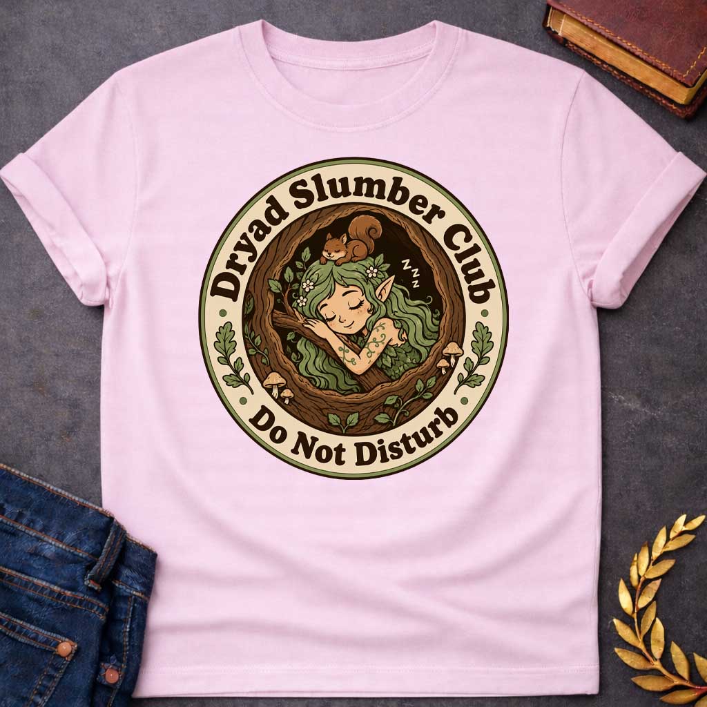Light Pink T-shirt with a cute, cartoon-style t-shirt graphic enclosed in a circular badge. Inside a cozy tree hollow, a little green-haired dryad or forest elf sleeps peacefully next to small mushrooms, while a tiny brown squirrel naps on her head. A cream-colored outer ring features dark brown text that reads "Dryad Slumber Club" along the top and "Do Not Disturb" along the bottom, accented by small green oak leaves