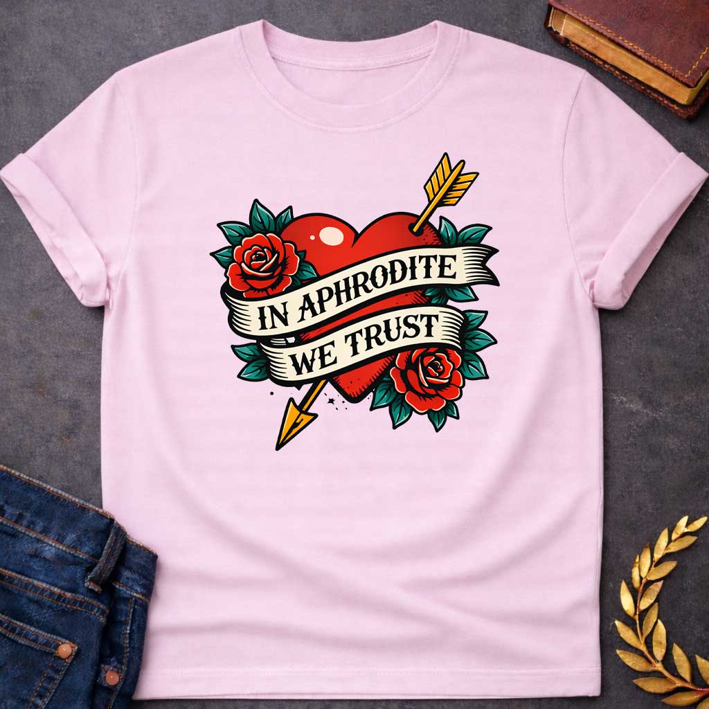 Light Pink T-shirt graphic in a classic American traditional tattoo style featuring a bold red heart pierced diagonally by a gold arrow, flanked by two red roses with teal green leaves, with a cream scroll banner wrapped around the heart reading "In Aphrodite We Trust" in black serif text — referencing Aphrodite, the Greek Goddess of Love, and Eros whose golden arrow inspired love