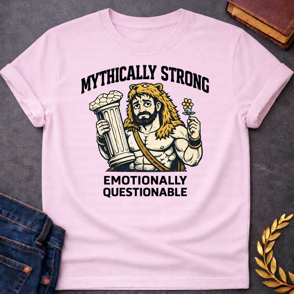 Light Pink T-shirt featuring a humorous illustration of a muscular, bearded man wearing a lion-skin hood—reminiscent of Hercules—with a sad, weary expression. He holds a broken classical column in one hand and a small flower in the other. Arched over the top is the white text "MYTHICALLY STRONG," and below the illustration is the text "EMOTIONALLY QUESTIONABLE,"