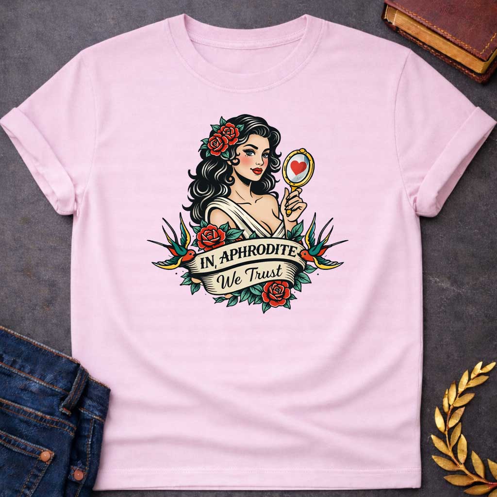 Light Pink T-shirt graphic in a classic American traditional tattoo style featuring a pin-up illustration of Aphrodite, the Greek Goddess of Love, with dark wavy hair adorned with red roses, wearing a white draped gown, holding a gold hand mirror reflecting a red heart. She is flanked by two swooping swallows and surrounded by red roses and green leaves, with a scroll banner below reading "In Aphrodite We Trust" in bold and script lettering