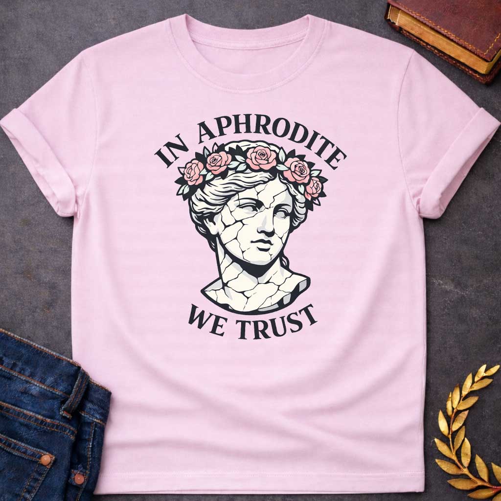 Light Pink T-shirt graphic featuring a cracked marble bust illustration of Aphrodite, the Greek Goddess of Love, adorned with a floral crown of pink roses and green leaves, with bold arched text reading "In Aphrodite" curved above and "We Trust" curved below, in a modern aesthetic vaporwave-inspired style with white text and soft pink and white tones