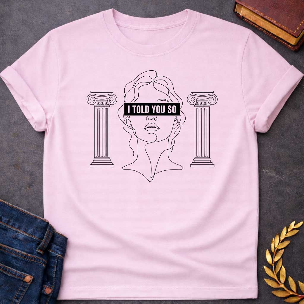 Light Pink T-shirt with a minimalist line art design featuring a continuous black ink sketch of Cassandra or a Greek goddess. Her eyes are covered by a bold, solid black rectangular bar with the text "I TOLD YOU SO" in white sans-serif capital letters. The portrait is flanked on both sides by simple, symmetrical line-drawn Ionic Greek columns.