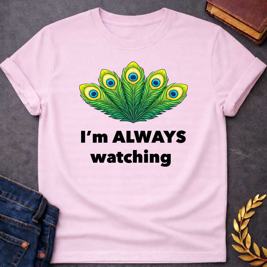 Light Pink T-shirt graphic featuring a vibrant illustrated fan of five peacock feathers in rich green, yellow, and blue tones, each with a distinctive eye-spot, above text reading "I'm Always Watching" — a humorous reference to Hera, the Greek Queen of the Gods, whose sacred symbol is the peacock