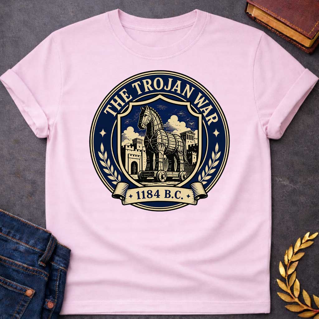 Light Pink T-shirt with a graphic featuring a formal vintage seal design in navy and cream. A detailed engraving-style illustration of the Trojan Horse on wheels is centered within a shield, set against the walled city of Troy with Greek temple columns and dramatic clouds. Text arched around the outer circle reads "The Trojan War" with gold diamond stars, flanked by cream laurel branches, and a scroll banner below reads "1184 B.C." — referencing the legendary Trojan War from Greek mythology