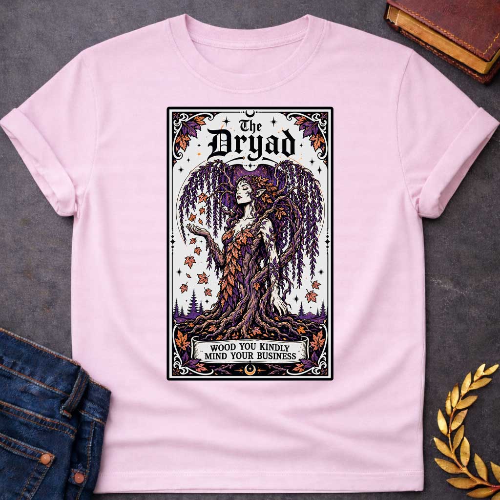 Light Pink T-shirt with a mystical, tarot-card style design framed by an ornate border. It features a majestic weeping willow dryad whose gown is woven from purple and orange autumn leaves. At the top, bold gothic text reads "The Dryad." A weathered banner at the base of her rooted trunk features a clever, pun-based tagline that reads: "WOOD YOU KINDLY MIND YOUR BUSINESS" in a clean, black serif font