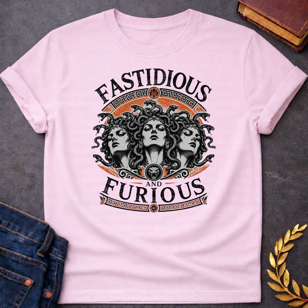 Light Pink T-shirt with a Humorous design combining 'Fast and Furious' with Greek mythology. Text reads 'FASTIDIOUS AND FURIOUS' in distressed lettering surrounding three faces of Medusa in black and white engraving style. Detailed serpents with individual scales form her hair. Greek key pattern borders in orange frame the composition with decorative sun motifs and a small snake head icon at center. Blends classical mythology with contemporary pop culture humor and clever wordplay.