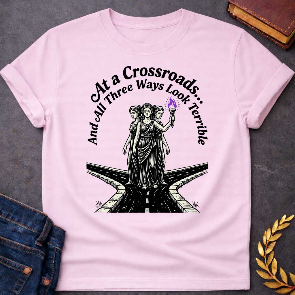 Light Pink T-shirt with an Illustration of the Hecate at a crossroads, rendered in black and white engraving style. The figures wear flowing robes and stand together, with the center holding a flaming torch with purple flames. Text reads 'At a Crossroads: And All Three Ways Look Terrible,' humorously referencing difficult choices and Hecate’s role as goddess of the crossroads. Multiple roads branch beneath them in perspective, receding into darkness symbolizing uncertainty.