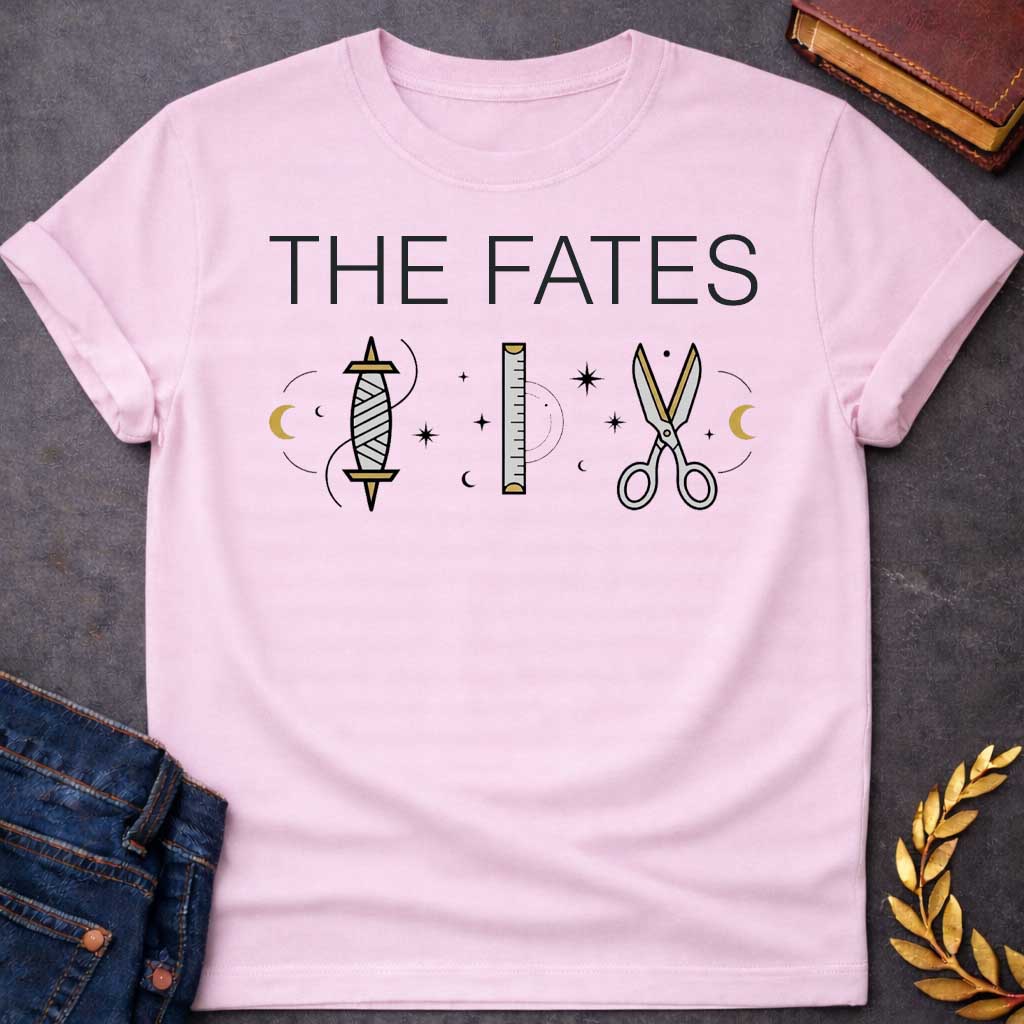 Light Pink T-shirt graphic featuring large bold black text reading "The Fates" above three minimalist icons representing the tools of the Moirai, the Greek Fates — a spindle with thread, a measuring ruler, and a pair of scissors — each surrounded by subtle crescent moons and celestial star details, rendered in white and gold tones
