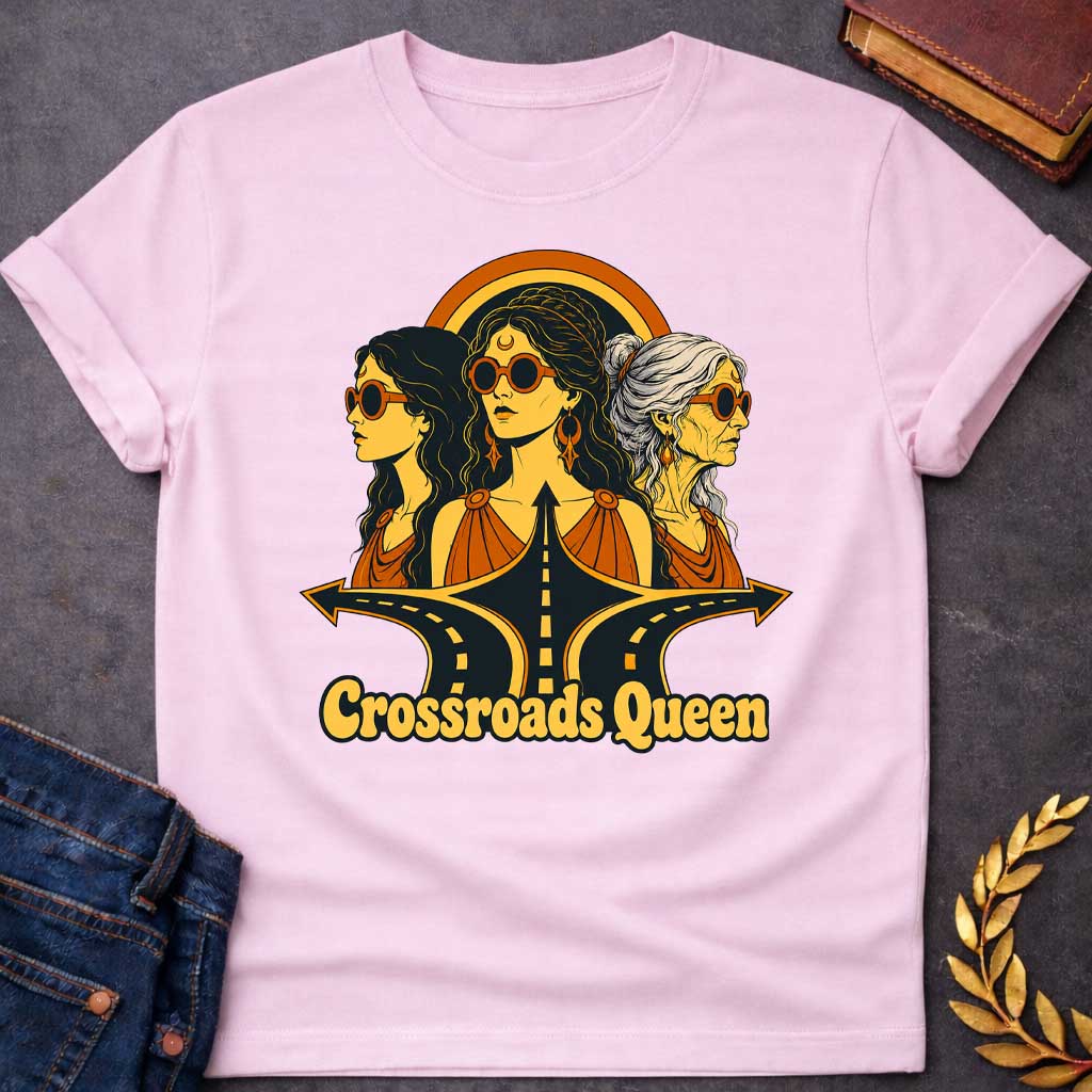 Light Pink T-shirt with a Glamorous retro illustration of Hecate as 'Crossroads Queen' in gold and orange with 70s aesthetic. Three female faces wear oversized round sunglasses and gold jewelry including crescent moon symbols and hoops. Center figure wears a crescent moon bindi. All wear draped orange clothing. A golden halo radiates behind them. Below, stylized black roads with yellow center lines converge at a crossroads. Text reads 'Crossroads Queen' in gold.