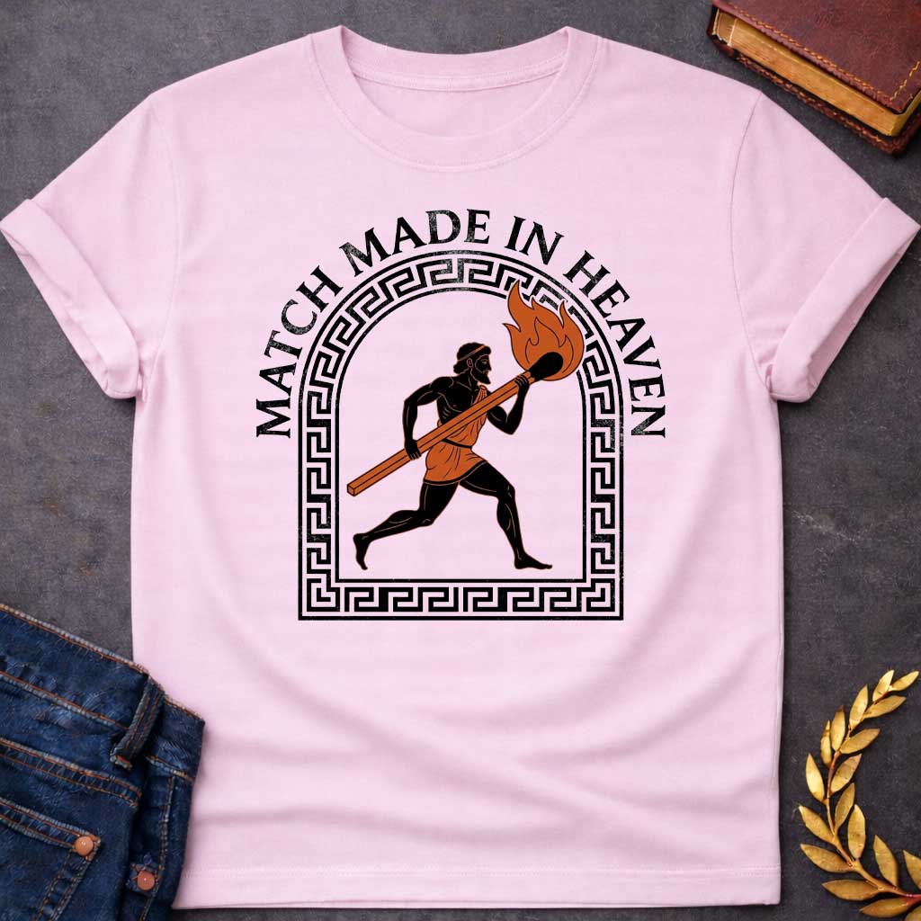 Light Pink T-shirt with A graphic in an ancient Greek Black-Figure pottery style featuring Prometheus running while carrying a massive, burning match stick. He is depicted in black silhouette with terracotta-orange details on his tunic and the flame. The figure is framed by a black Greek key meander arch. Above the design, the pun "MATCH MADE IN HEAVEN" is written in an arched, bold black serif font.