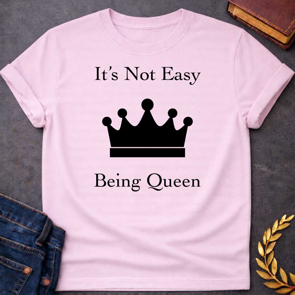 Light Pink T-shirt graphic in white on black featuring elegant serif text reading "It's Not Easy Being Queen" split above and below a simple white crown silhouette icon, in a humorous nod to Hera, the Greek Queen of the Gods