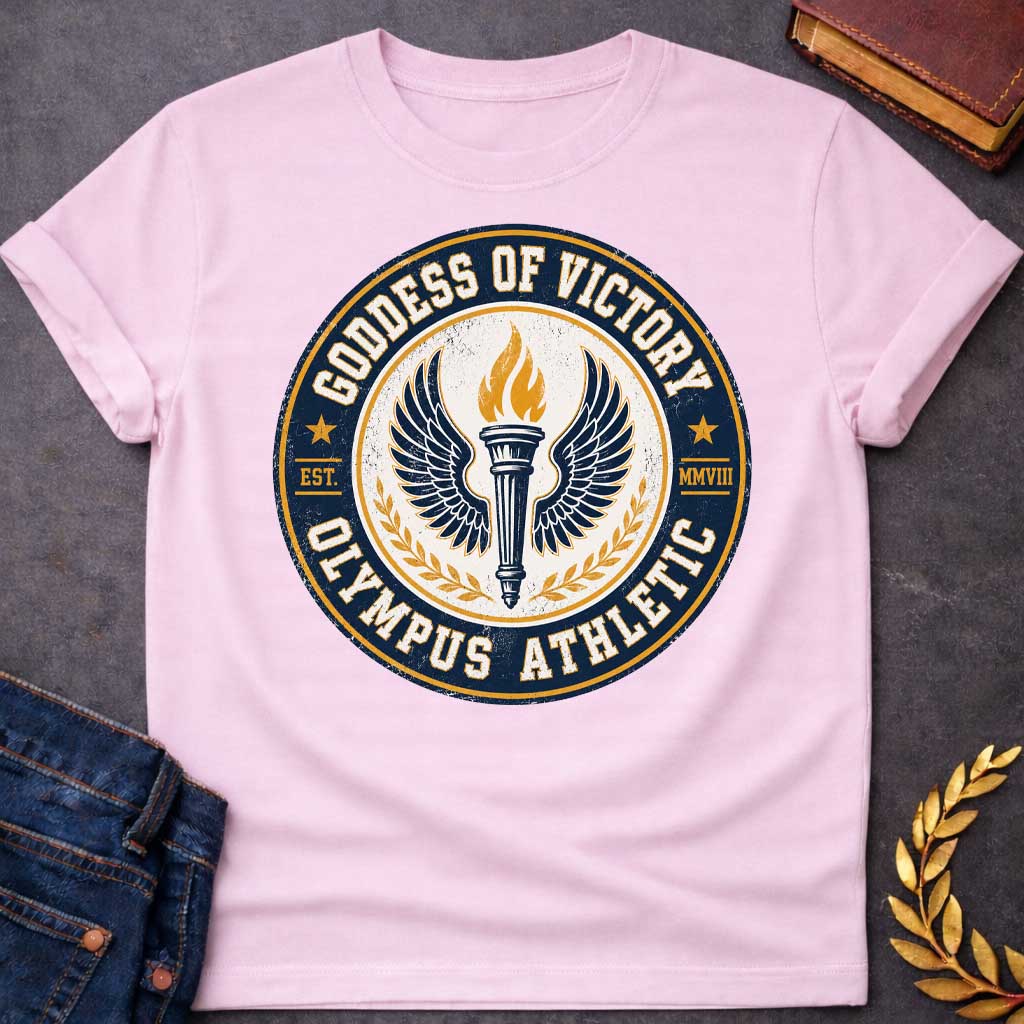 Light Pink T-shirt with an Official athletic seal in navy blue, cream, and gold featuring a torch with large outstretched wings symbolizing Nike, the goddess of victory. Flames burn from the torch. Olive wreaths frame either side. Text reads 'GODDESS OF VICTORY' and 'OLYMPUS ATHLETIC' around the circular border with 'EST. MMVIII.' Distressed vintage texture and gold stars enhance the emblem. Celebrates athletic achievement and triumph.