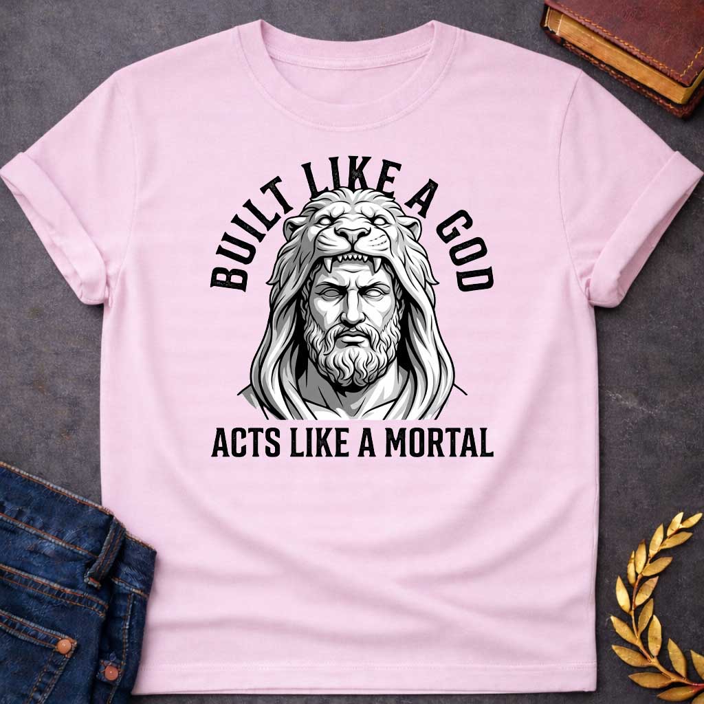 Link Pink T-Shirt with a high-contrast, black-and-white graphic. The illustration depicts a stylized, statuesque man wearing the head and mane of a lion as a hood, reminiscent of Hercules. The text "BUILT LIKE A GOD" is arched over the top, and "ACTS LIKE A MORTAL" is printed in bold, straight text across the botto