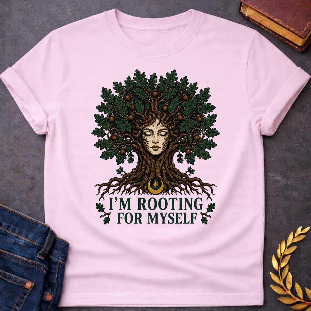 Light Pink T-shirt with a cottagecore-style graphic featuring a detailed illustration of an oak tree dryad or nature spirit. Her serene, closed-eyed face forms the center of the tree trunk, with gnarled roots expanding below and a massive canopy of dark green oak leaves and brown acorns spreading above. Underneath the roots, a pun-based tagline reads "I'M ROOTING FOR MYSELF" in a bold, clean green serif font.