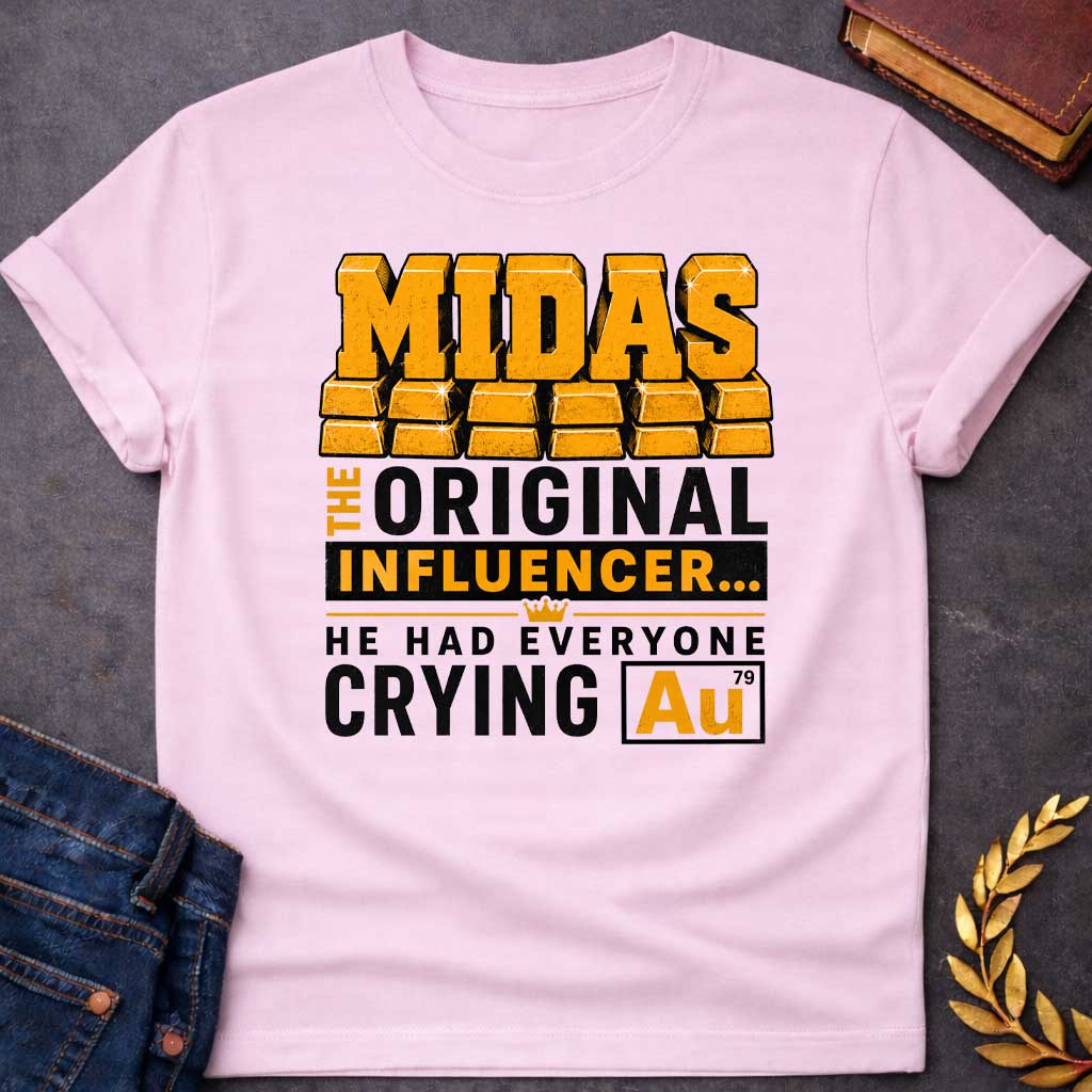 Light Pink t-shirt with A Bold typographic Greek mythology design featuring stacked gold bar lettering spelling "MIDAS" above the phrase "The Original Influencer... He Had Everyone Crying Au." The design uses distressed black and metallic gold typography with a small crown icon and a gold-themed chemical element "Au" symbol, creating a humorous King Midas pun in a vintage-inspired graphic style.
