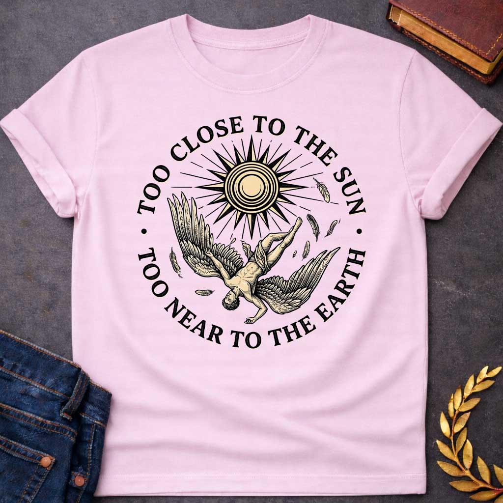 Light Pink t-shirt with a vintage engraved-style illustration. A winged figure of Icarus plummets downward upside down as loose feathers drift through the air beneath a large, radiating sun emblem. Encircling the detailed central artwork, bold black serif text reads "TOO CLOSE TO THE SUN" along the top arch and "TOO NEAR TO THE EARTH" along the bottom curve.