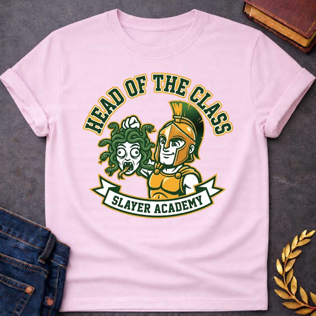 Light Pink t-shirt in collegiate mascot style: a smirking gold-helmeted Greek warrior holds up Medusa's shocked, snarling severed head by her green snake hair. Bold green and gold text arcs above the design, with "Slayer Academy" on a white ribbon banner below. Playful varsity-logo aesthetic on a white background.