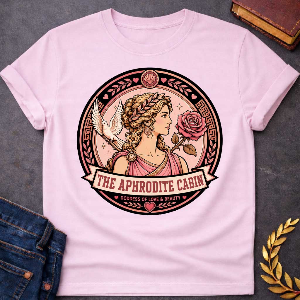 Light Pink t-shirt with a Aphrodite Greek mythology cabin design featuring the goddess of love and beauty in a pink robe and laurel crown, surrounded by a rose, white dove, hearts, seashells, and Greek key details. Circular pink and black emblem includes the text “The Aphrodite Cabin” and “Goddess of Love & Beauty,”